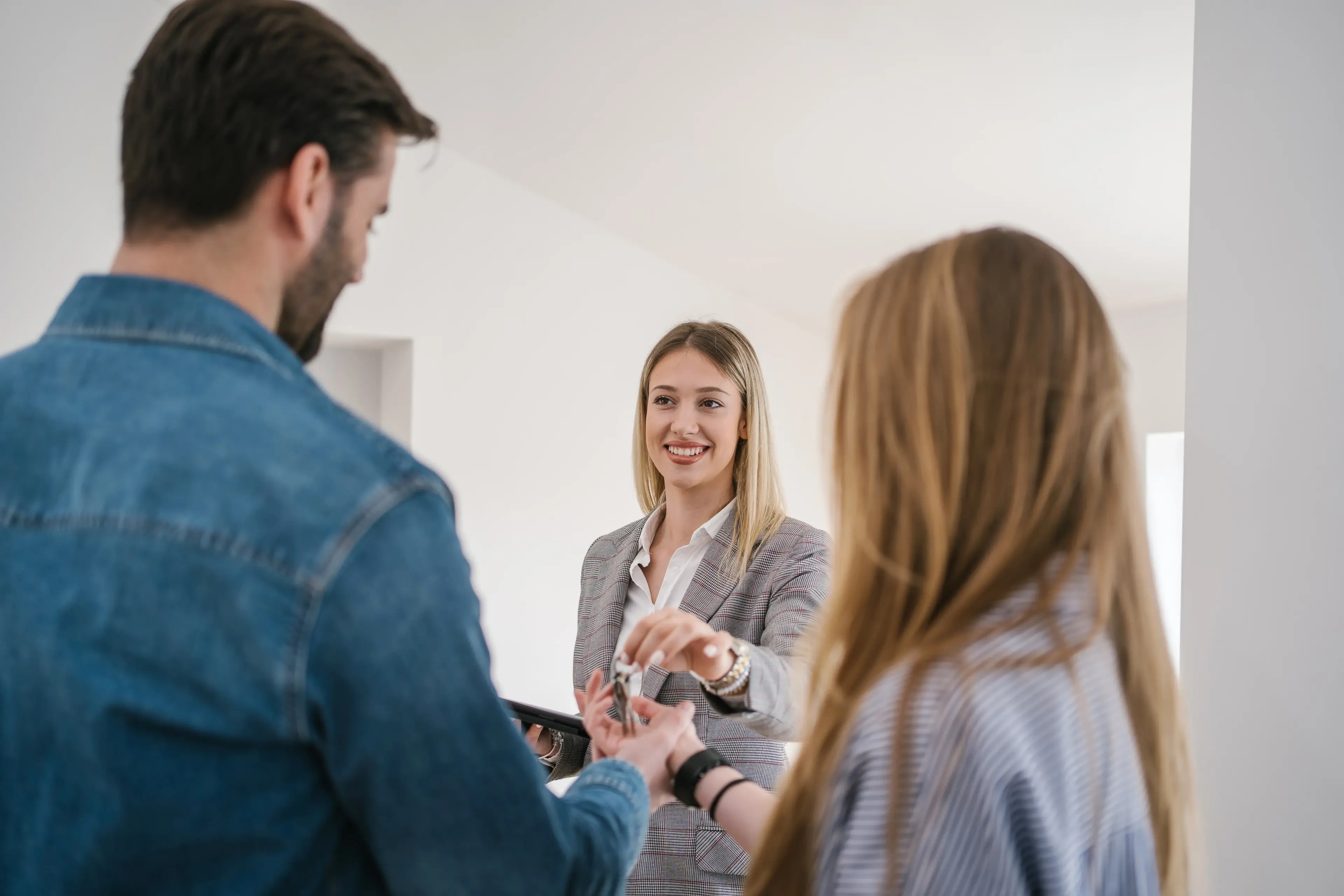 Immobilienmaklerin übergibt Schlüssel an ein Paar in ihrem neuen Zuhause. Eine professionelle Frau im karierten Blazer lächelt, während sie in einem hellen, leeren Raum Hausschlüssel an ein von hinten zu sehendes Paar übergibt.