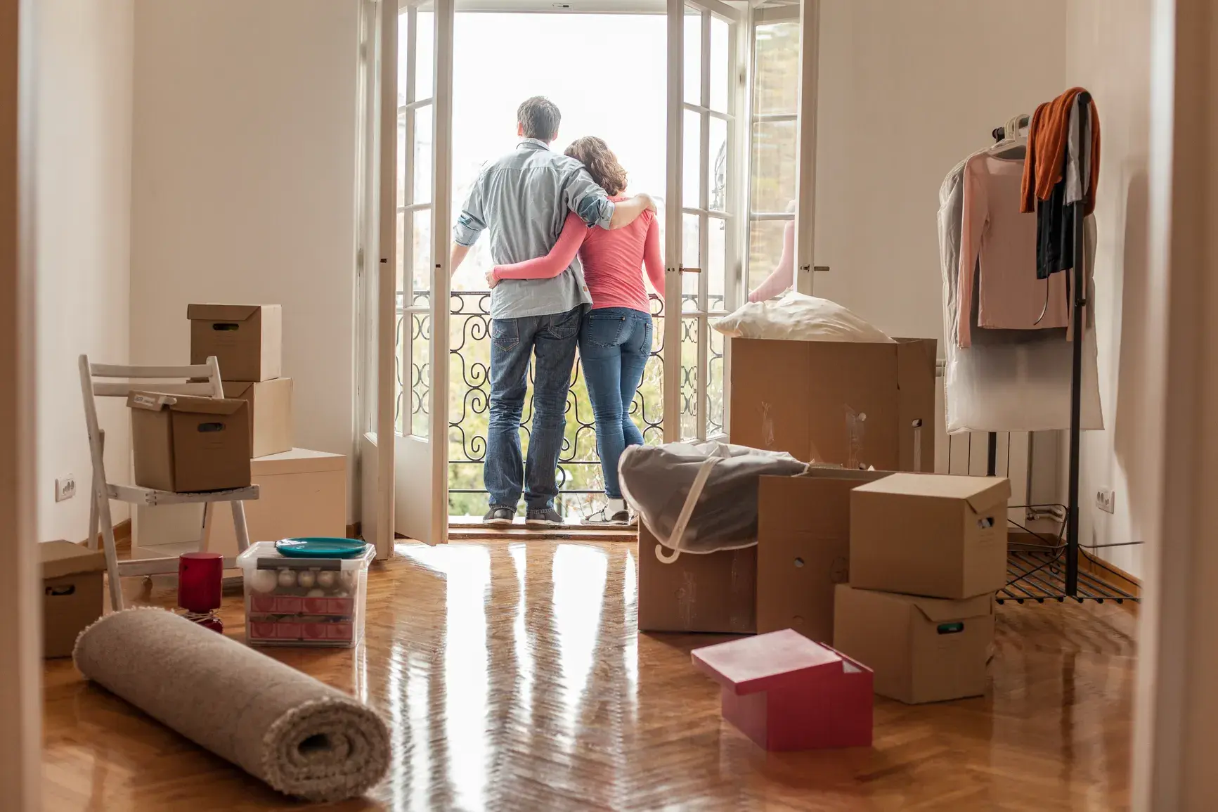 Paar blickt von Balkontüren in neuer Wohnung umgeben von Umzugskartons nach draußen. Rückansicht eines sich umarmenden Mannes und einer Frau, die durch offene französische Türen schauen, mit ausgepackten Kartons, einem aufgerollten Teppich und einem Kleiderständer in einem hellen Raum mit Holzböden.